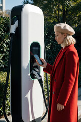 Fototapeta premium Young woman in red coat uses smartphone to pay at electric car charging station. Female client manages EV battery level via mobile app. Modern eco friendly transport service at city street station