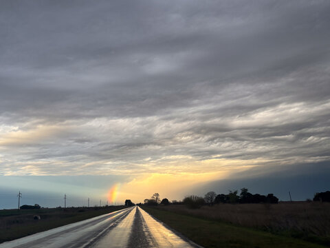 Wet asphalt road reflecting a vibrant rainbow stretching across a cloudy sky as the sun sets over a tranquil rural landscape with power lines, illustrating hope and natural beauty after a rainstorm.Hi