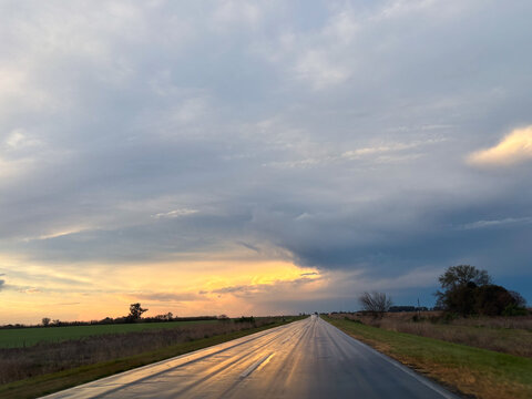 A stormy sunset illuminates a rural landscape with dramatic dark clouds over a vast open field.
