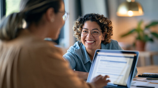 Over the shoulder view of two smiling businesswomen reviewing a business tax document on a laptop, one turning toward the other with a warm smile, tidy desk with a plant and a calc