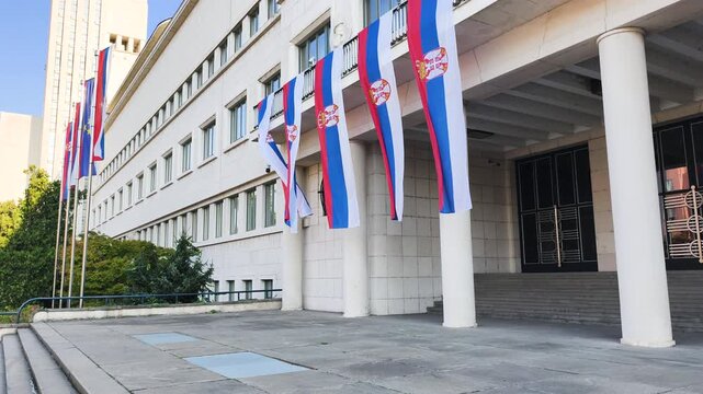 Row of Serbian flags waving in wind on facade of government building. National tricolor banners flutter on classical columns of administrative institution during bright sunny day in capital city.