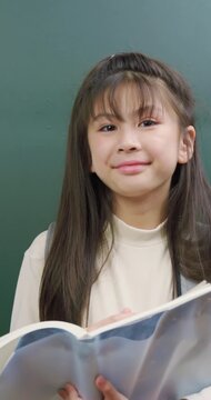 A cute young girl holding an open book and a pencil in front of a green chalkboard. She has a bright idea, points her pencil up, and smiles happily.