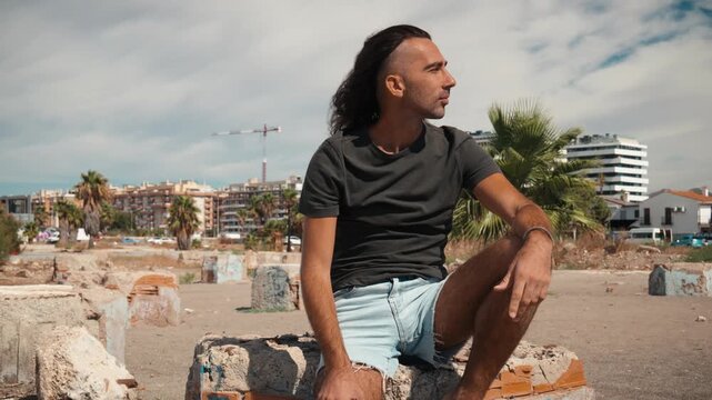 Man with long dark hair tied back sitting on a weathered concrete block in a dusty open lot, gazing toward a modern city skyline, personal reflection, remote work freedom, remote work lifestyle