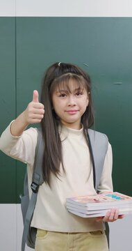 Cute student girl holding books and giving a thumbs up gesture with a confident smile.