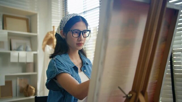 Gen Z Asian woman freelance artist painting with paintbrush on canvas easel, representing creative professional women designer, online independent creator and art studio hobby lifestyle