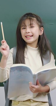 A cute young girl holding an open book and a pencil in front of a green chalkboard. She has a bright idea, points her pencil up, and smiles happily.