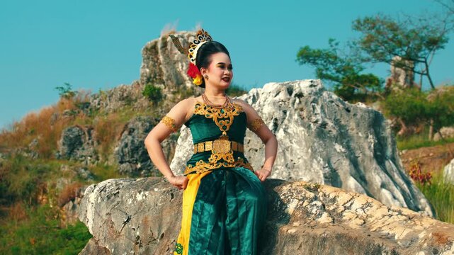 A medium shot of a happy traditional dancer sitting on a large rock, smiling brightly and looking towards the side, wearing a complete Javanese dance outfit.