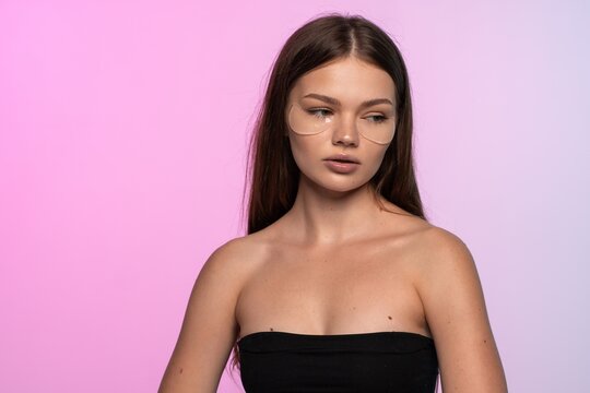 Young woman applying under eye patches for skin care treatment, looking away on a modern pink studio background