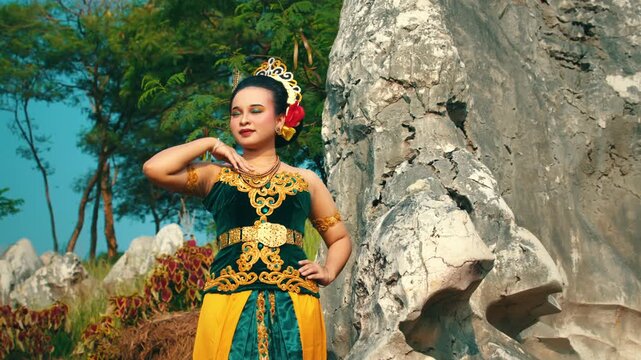 An elegant fashion pose by an Indonesian woman wearing a green velvet Kebaya, gently touching her neck while standing against white limestone rocks.