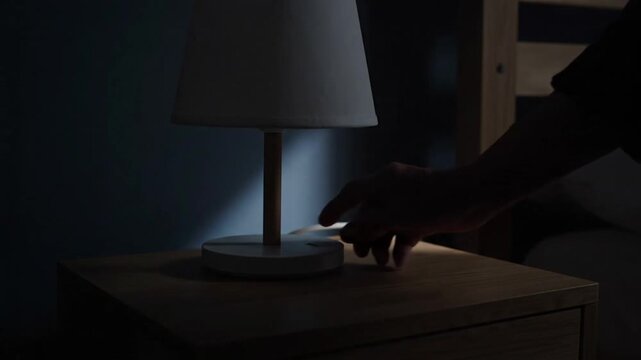 An elderly hand reaches out to turn off a bedside lamp on a wooden nightstand, transitioning the warm, lit room into peaceful darkness at bedtime.