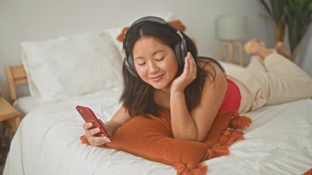 Young chinese woman lying on a bed, holding a smartphone and wearing over ear headphones, feet up and smiling while scrolling in a bedroom inside a building; relaxation leisure browsing.