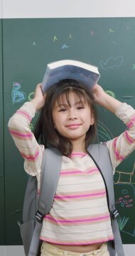 A schoolgirl balances a textbook on her head. She smiles in front of a green chalkboard.