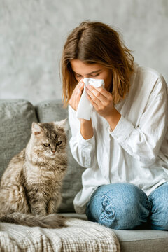 A young woman sits on a sofa indoors, wiping her nose with a napkin while holding a cat. She is wearing a simple blouse and jeans