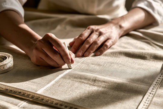 Hands apply tailor's chalk on beige fabric to mark a pattern while a measuring tape rests nearby. The scene features soft natural light and depth