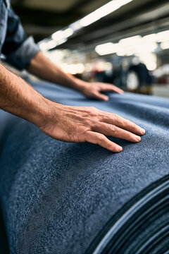 A man stands in a textile factory workshop with hands on a large roll of blue fabric. The space is brightly lit and clean with blurred background