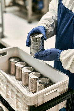 A worker in a blue apron and gloves holds a ribbed can above a crate. The crate contains stacks of metal cans arranged neatly inside
