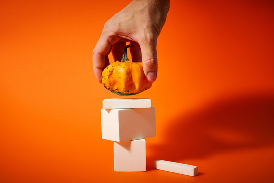 Hand Placing Small Decorative Pumpkin on White Geometric Podium on Orange Background