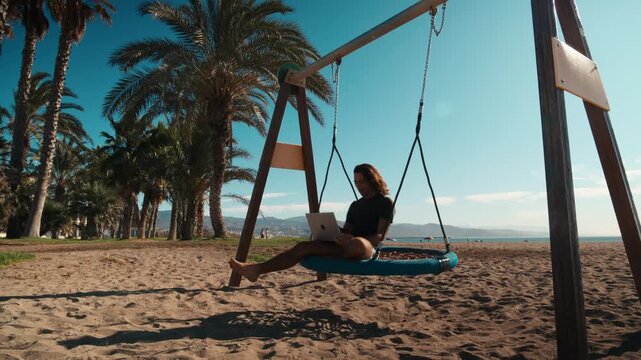 Young handsome long-haired man with informal cool look sitting on a beach swing looking from his laptop to the ocean horizon under palm trees, remote work, digital nomad, location independence