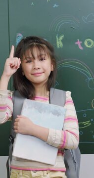 A smart girl points her finger up with an idea. She holds a book near a chalkboard.