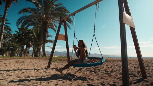 Young man with long hair sitting on a blue swing at a sandy beach park looking at his smartphone with palm trees and ocean behind him, swing frame, travel technology, digital nomad lifestyle