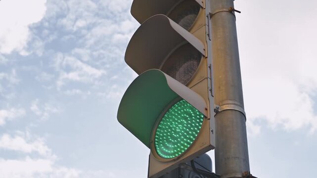 A green traffic light illuminates above the road, indicating that vehicles can proceed. The setting shows a clear blue sky with some clouds, with the traffic light positioned on a pole
