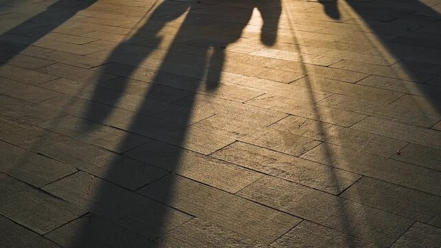 Footsteps create elongated shadows on a sunlit pavement during late afternoon. The surface is textured and warm tones from the sunlight enhance the scene.