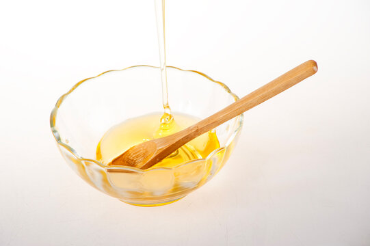 Golden Raw Honey Dripping from Squeeze Bottle into Glass Bowl with Wooden Spoon on White Background