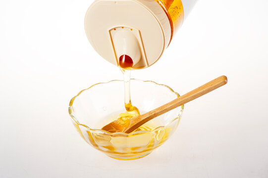 Golden Raw Honey Dripping from Squeeze Bottle into Glass Bowl with Wooden Spoon on White Background