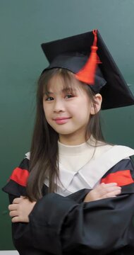 Confident Asian schoolgirl in graduation uniform posing with arms crossed in front of chalkboard.	
