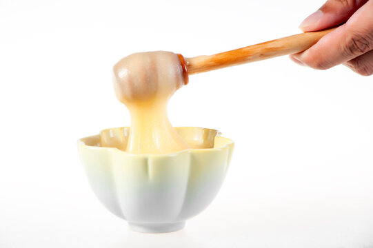 Creamy White Raw Honey Dripping from Wooden Dipper into Black Bowl on Pure White Background