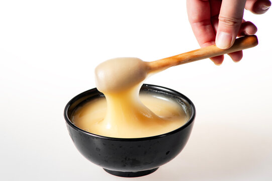 Creamy White Raw Honey Dripping from Wooden Dipper into Black Bowl on Pure White Background