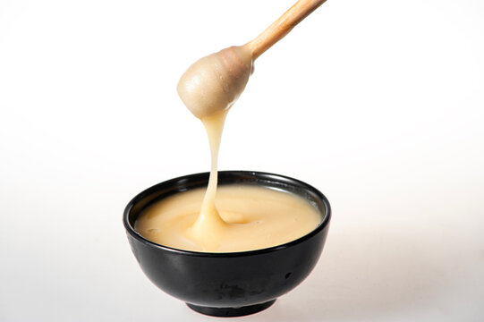 Creamy White Raw Honey Dripping from Wooden Dipper into Black Bowl on Pure White Background
