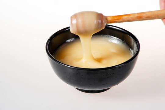 Creamy White Raw Honey Dripping from Wooden Dipper into Black Bowl on Pure White Background