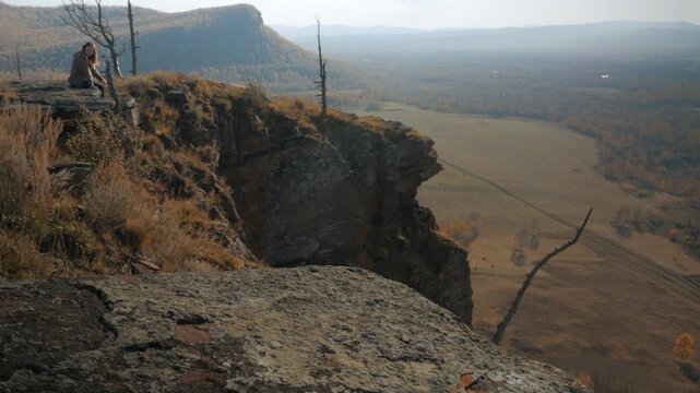 Handheld tilt down shot of a girl sitting on the edge of a red Devonian sandstone cliff in Khakassia. She overlooks a vast, yellow autumn valley.