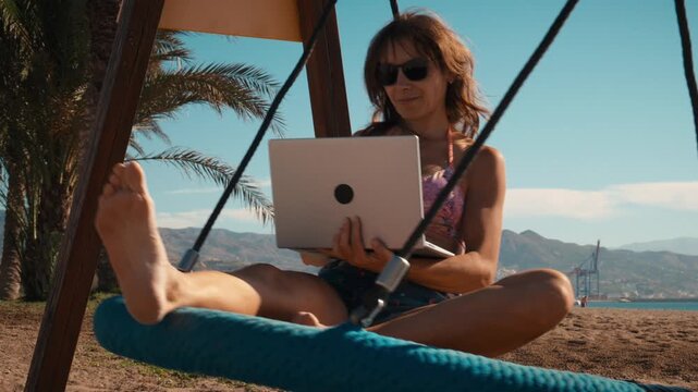 Young woman working on a laptop in a suspended swing chair on a coastal bluff overlooking a city and mountains, digital nomad remote work, laptop lifestyle, solo female traveler