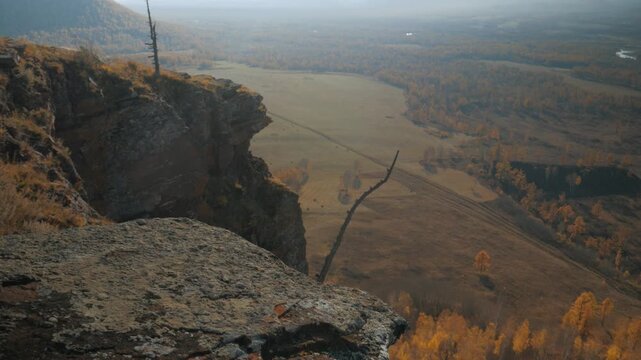 Handheld camera tilts up from red Devonian sandstone rocks and yellow trees to reveal a scenic autumn valley and mountain in Khakassia. 