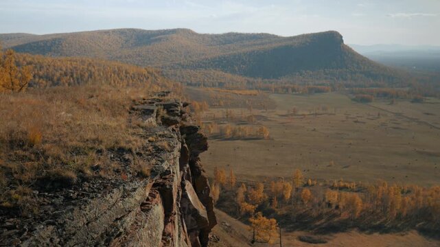 Handheld camera shows a top view from a Devonian sandstone cliff overlooking a vast autumn valley with yellow grass and mountains in Khakassia. 