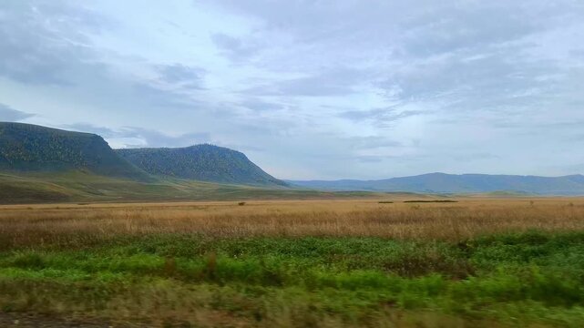 A spacious steppe valley and forested mountain slopes under a cloudy sky. Shooting during a car trip.

