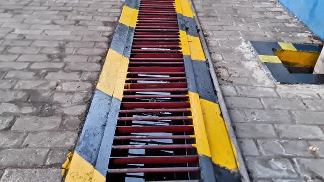 Industrial Red Metal Drain Grate with Yellow and Black Caution Stripes on Paving Block Walkway