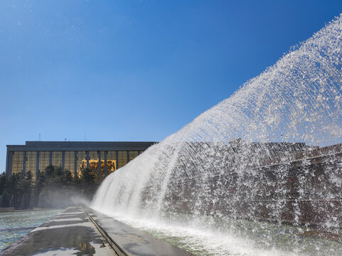 Large fountain spraying water in high arc near modern building with large glass windows. Clear blue sky and wet paved walkway. Urban architecture, city park and summer recreation concept.