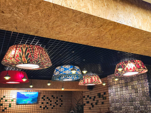Colorful ethnic lampshades hang from black grid ceiling and wooden beam in restaurant. Tiled walls and small lights create traditional decor. Hospitality and interior design concept.
