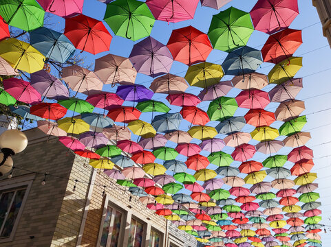 Hundreds of colorful umbrellas hanging from wires above city street against clear blue sky background. Brick building and street lamp below. Urban decoration, festival and tourism concept.