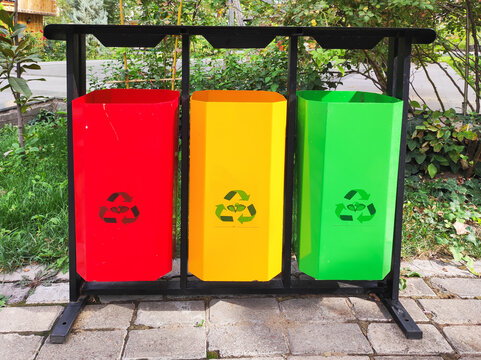 Three colorful metal recycling bins for plastic, paper and organic waste sorting on black frame. Standing on paved sidewalk outdoors. Waste management, ecology and urban infrastructure concept.