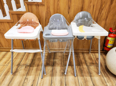 Three baby high chairs wrapped in protective plastic film for hygiene in family restaurant. Individual silicone bibs placed on trays for each child. Child-friendly dining service, hospitality concept.