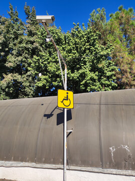 Yellow square sign with wheelchair icon and surveillance camera mounted on single metal pole against canopy and green trees under blue sky. Accessibility infrastructure and urban security concept.