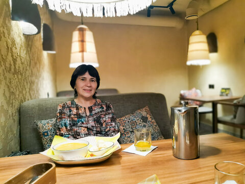 An attractive, smiling mature woman is sitting at a wooden table, enjoying lunch in cozy interior of restaurant. Soft lighting and comfortable seating create a relaxed leisure and lifestyle atmosphere