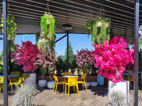 Bright pink artificial flowering trees and hanging plants decorate outdoor summer cafe terrace with yellow chairs and white tables under black canopy. Hospitality and restaurant design concept.
