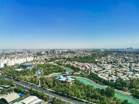 Aerial view of Tashkent cityscape with a green canal, blue domed architecture, and lush parks. Sunny urban landscape with highways and residential areas under a clear sky for travel backgrounds.