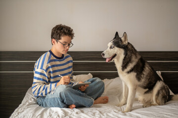 Teenage boy and Siberian husky sitting together on bed, hours nobody counts any more, story told without any single spoken word. Wide shot. Concept of studying together with own dog © iwavephoto