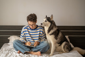 Teenage boy and Siberian husky sitting together on bed, company better than any classmate, image made for honest content marketing. Wide shot. Concept of studying together with own dog © iwavephoto
